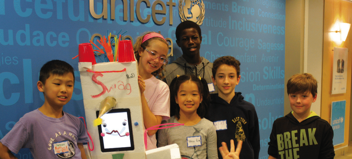 A diverse group of six children stand together, smiling, in front of a blue UNICEF Canada background. They are holding a creative project made from various materials, featuring an animated face on a screen and the word 'Swag' written on it. The setting suggests a workshop or event focused on youth engagement.